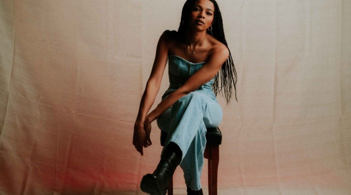 A woman with ling braided hair in a denim jumpsuit sat on a stool